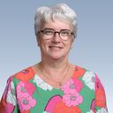 A smiling woman with short white hair and glasses, wearing a colorful floral blouse, against a blue gradient background.